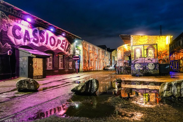 Leuchtend beleuchtete, graffiti-bedeckte Clubfassade mit "Cassiopeia" Schriftzug links, große Steine auf nassem Boden, Pfützen spiegeln das Licht, dunkler Abendhimmel im Hintergrund.