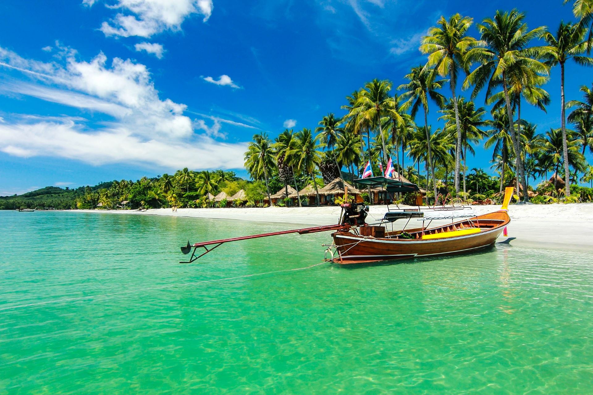 Typisches thailändisches Longtail-Boot schwimmt auf türkisblauem Wasser nahe einem weißen Sandstrand mit hohen Palmen unter einem strahlend blauen Himmel.