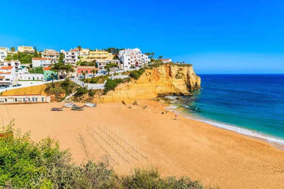 Strand mit goldenem Sand und blauen Wellen. Links bunte Häuser auf einer Klippe, vorne ein paar Boote am Strand. Klare, leuchtende Farben unter strahlend blauem Himmel.