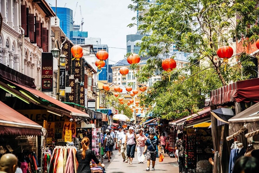 Straße eines chinesisch geprägten Viertels. Rote Lampions hängen über der Strasse und zahlreiche Stände stellen ihre Waren auf der Strasse aus.