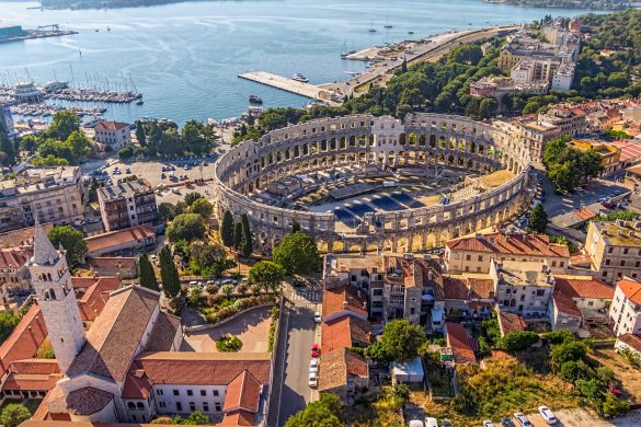 Vogelansicht auf Pula. Alte Steingebäude reihen sich um die Überreste eines runden Gebäudes. Im Hintergrund das Meer.