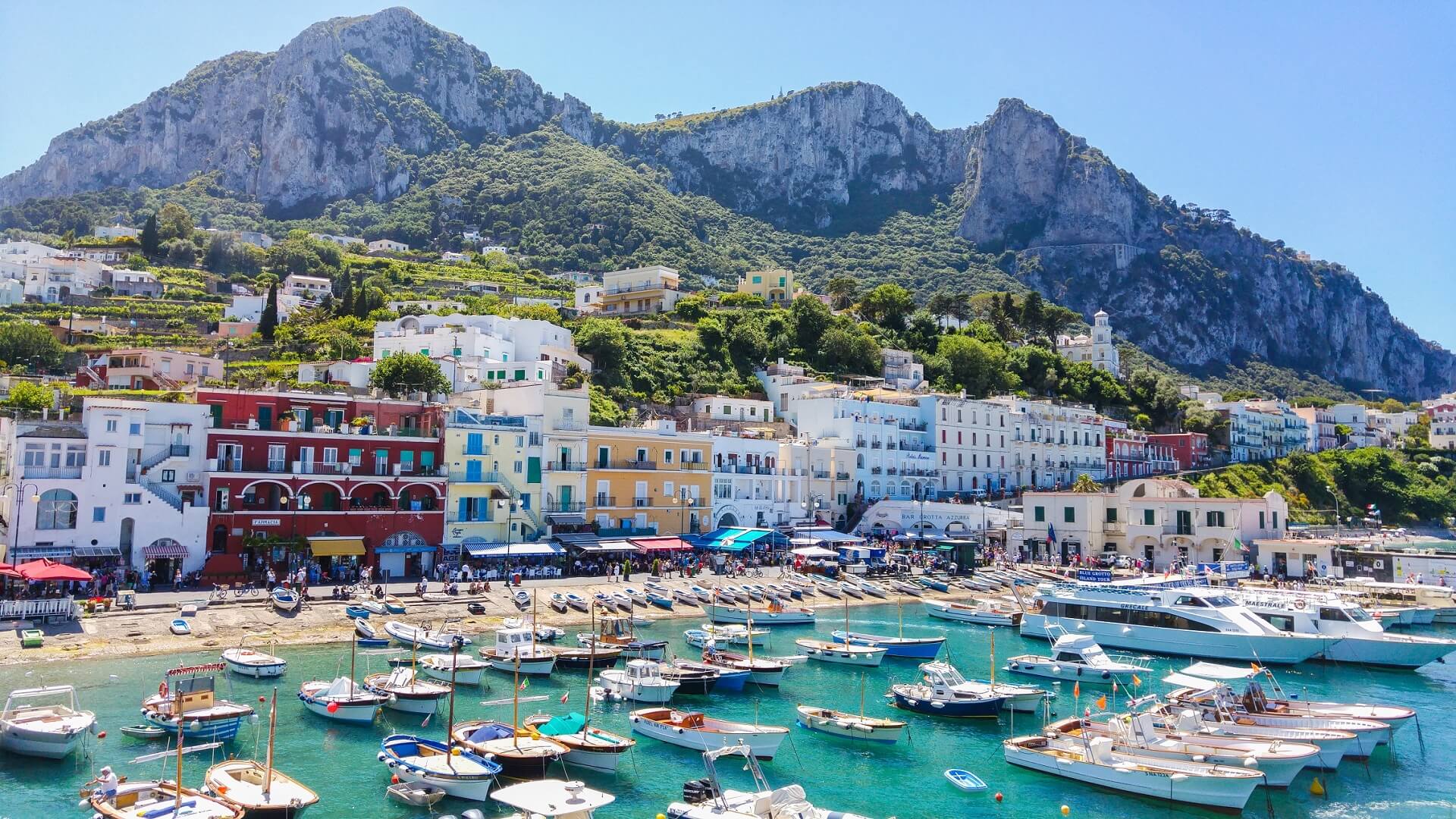 Bunte Häuser reihen sich am Hafen von Marina Grande, Capri. Im Vordergrund schwimmen Boote im türkisblauen Wasser, während saftig grüne Hügel und Felsen die Kulisse bilden, bei strahlendem Sonnenschein.