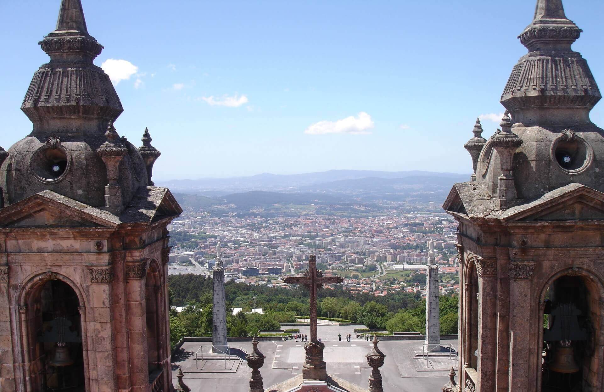 Im Vordergrund zwei historische Türme, in der Mitte ein Kreuz. Im HIntergrund sieht man den Ausblick auf die Stadt.