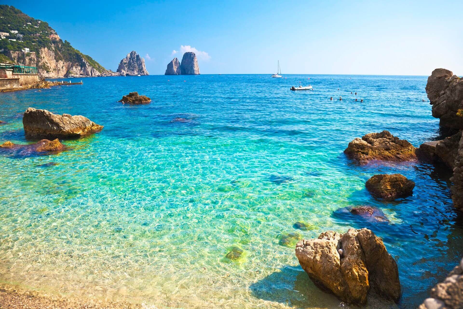Klares, türkisfarbenes Wasser an Capris Küste. Im Vordergrund liegen große Felsen; im Hintergrund stehen markante Faraglioni-Felsen im Meer. Am Horizont segelt ein Boot unter klarem Himmel.