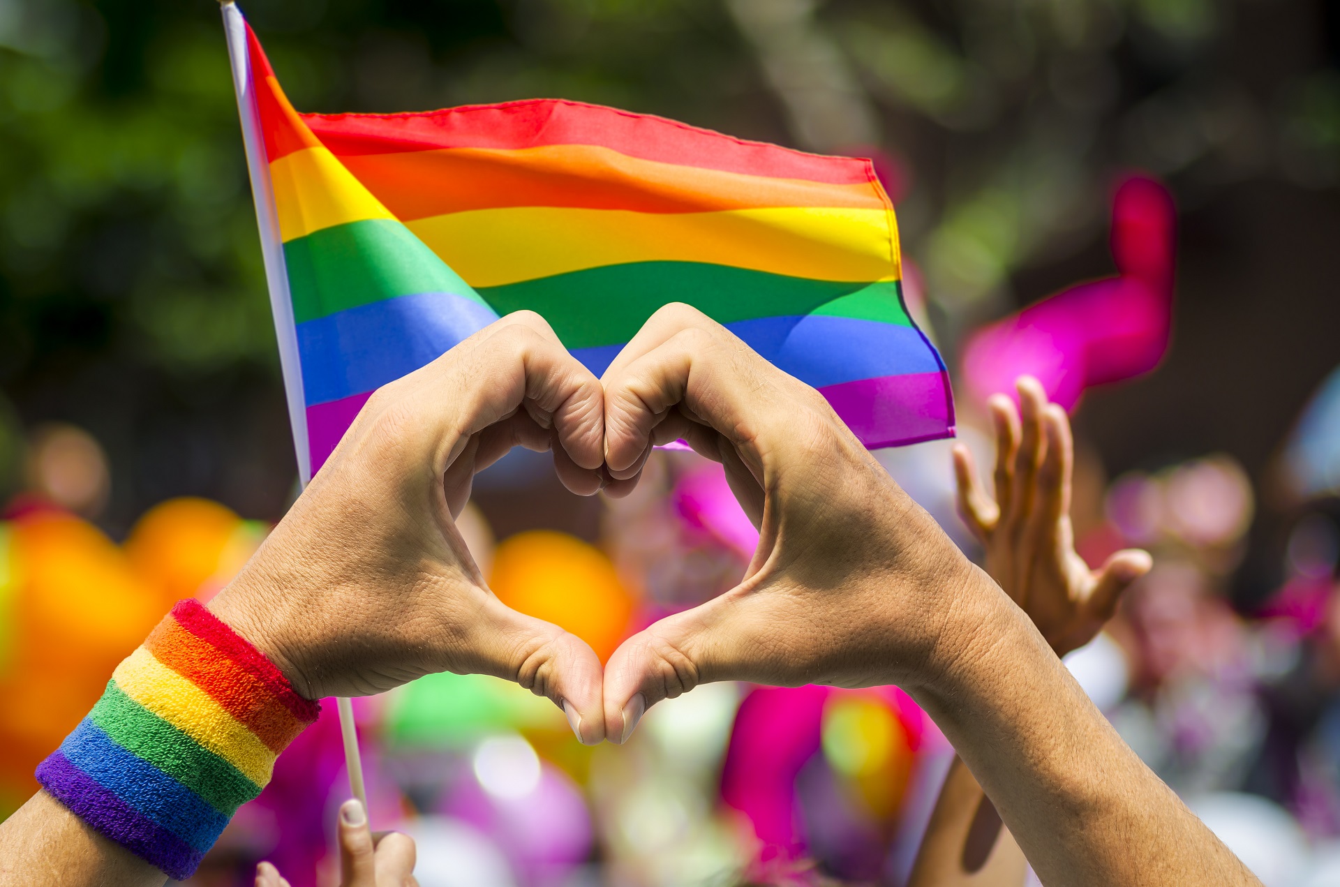 Zwei Hände bilden ein Herz vor einer Regenbogen Fahne