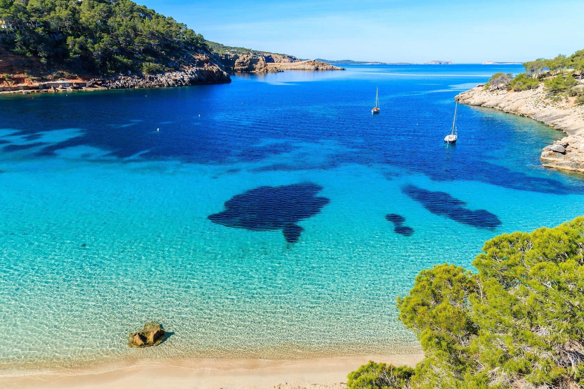 Klares türkisblaues Wasser einer Bucht mit schwimmenden Menschen. Links Boote, rechts goldene Felsen und dichter Kiefernwald, im Hintergrund ein belebter Sandstrand mit zahlreichen Badegästen.