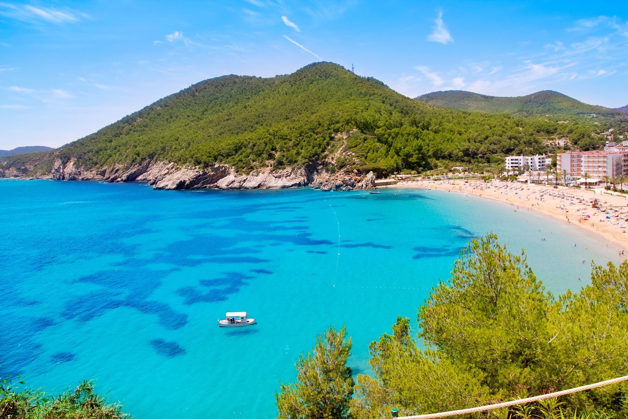 Klares, türkisfarbenes Wasser mit einem Boot in der Mitte; links felsige Küste, dahinter bewaldete Hügel; rechts breiter Sandstrand mit Besuchern und weißen Gebäuden im Hintergrund; blauer Himmel.
