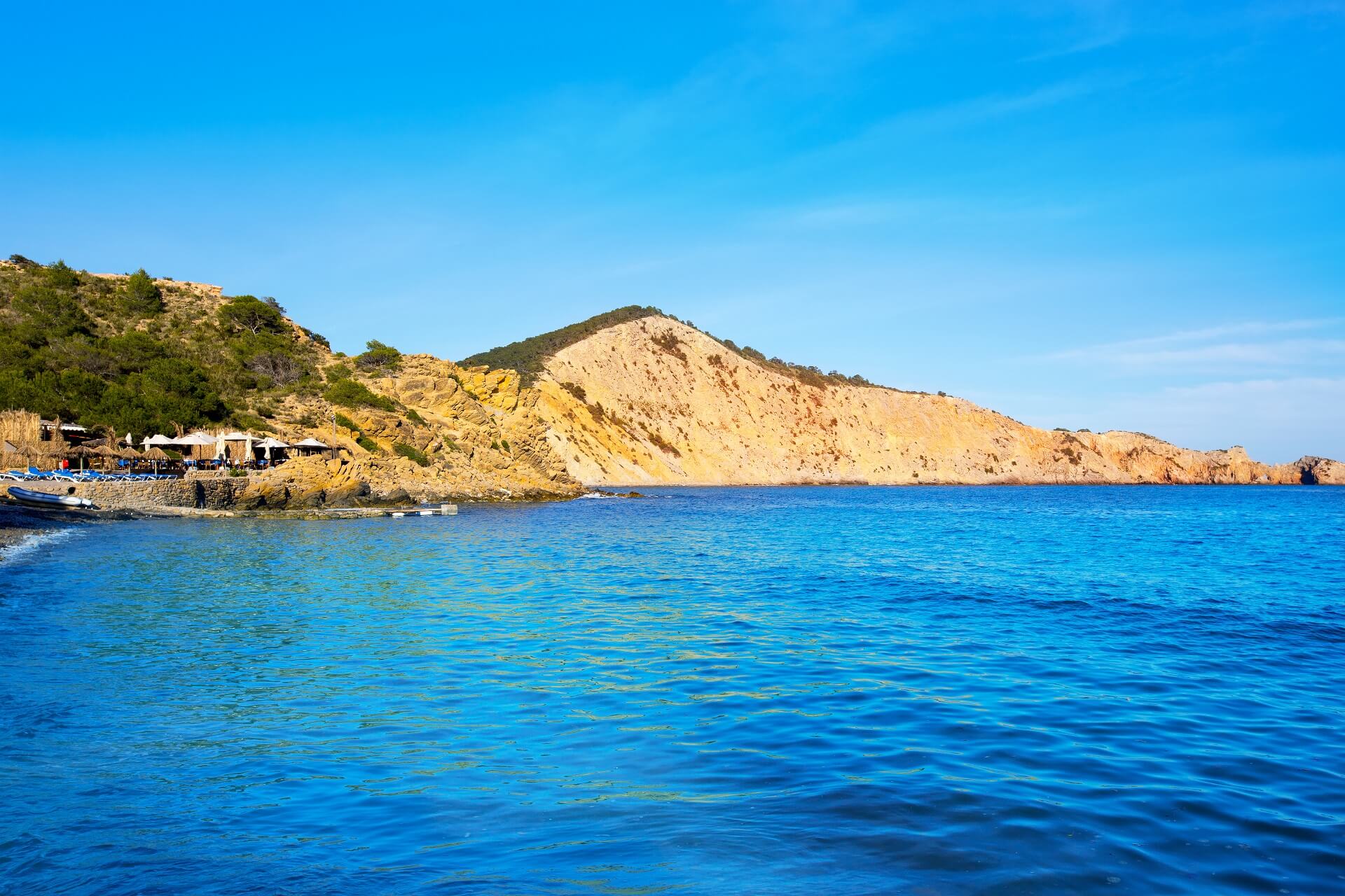 Klares blaues Meer im Vordergrund, links eine felsige Küste mit grüner Vegetation und einem Restaurant. Ein sandfarbener Hügel erstreckt sich im Hintergrund unter blauem Himmel.
