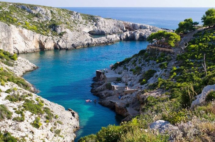 Eine ruhige, türkisfarbene Bucht mit klarem Wasser, umgeben von felsigen grünen Klippen. Menschen schwimmen und entspannen auf den Felsen. Rechts befindet sich ein Pfad mit Bäumen.