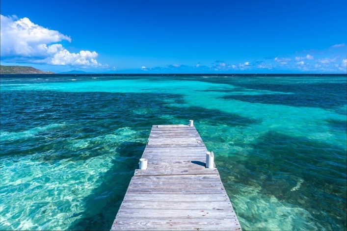 Ein hölzerner Steg führt mittig ins türkisblaue karibische Meer. Links ist eine Hügelküste zu sehen. Der Himmel ist strahlend blau mit ein paar weißen Wolken am Horizont.