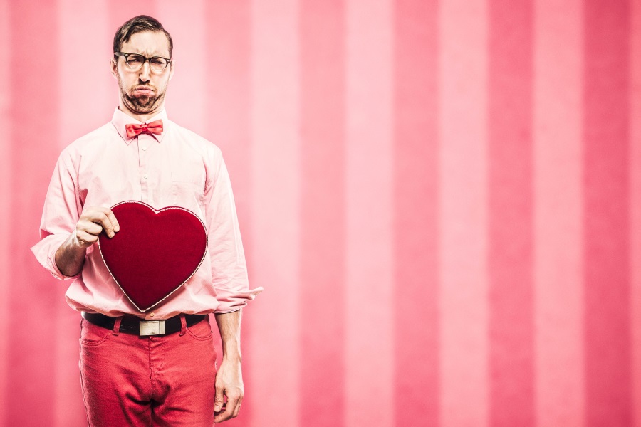 Ein missmutiger Mann in rosa Kleidung hält widerwillig eine herzförmige Schachtel, im Hintergrund pink-weiße Streifen, verkörpert einen Valentinstag-Hasser.