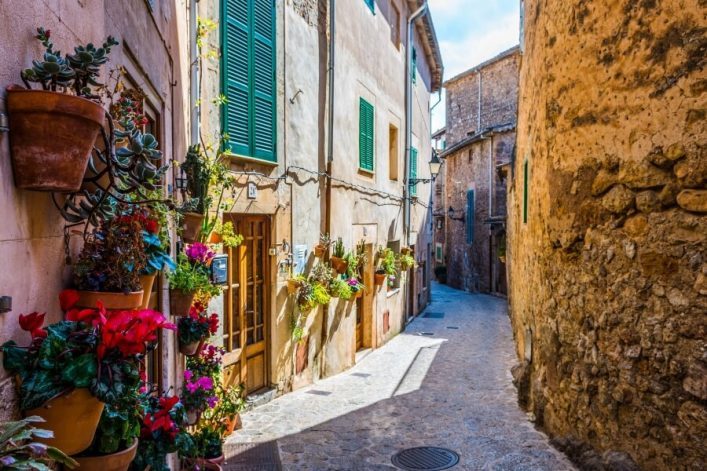 Enge, gepflasterte Gasse in Valldemossa mit blumengeschmückten Häusern. Links: helle Wände, grüne Fensterläden, bunte Pflanzen in Töpfen. Rechts: natursteinfarbene Wand, Sonnenlicht erhellt den Weg.