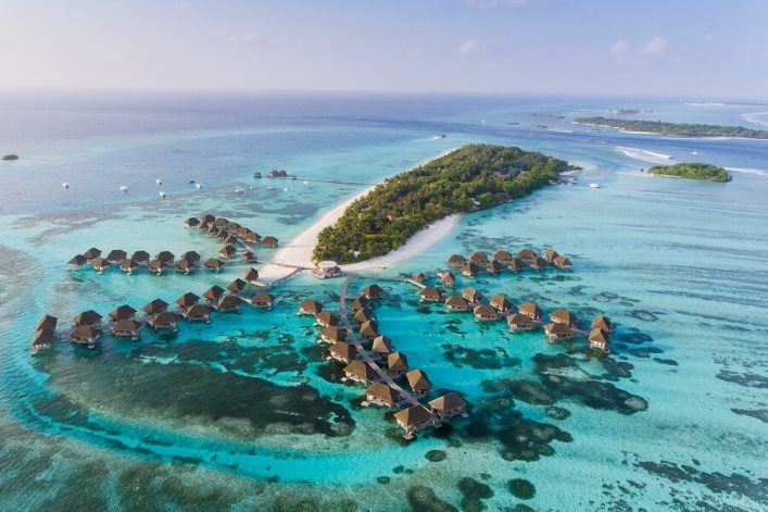 Wasservillen auf türkisblauem Meer umgeben eine grüne Insel mit weißem Sandstrand in der Mitte. Ein langer Steg verbindet die Villen mit der Insel. Boote sind im Wasser verstreut.