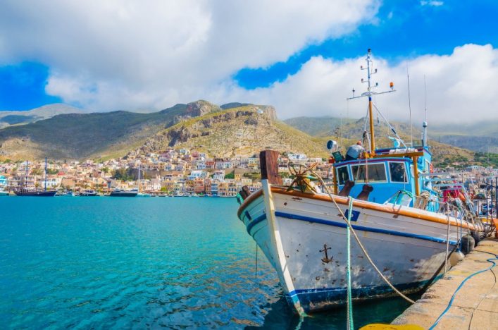 Ein weiß-blaues Fischerboot liegt im Vordergrund am Pier. Im Hintergrund erstreckt sich ein malerisches Dorf mit bunten Häusern vor einer bergigen Landschaft unter bewölktem Himmel.