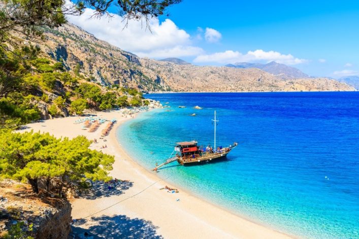 Touristenboot am klaren Wasser der Apella-Bucht auf Karpathos, Griechenland. Der Strand liegt links, umgeben von grünen Bäumen und sanften Hügeln. Rechts strahlt das tiefblaue Meer.