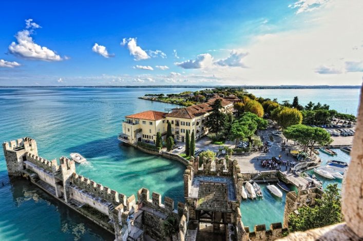Festung mit Zinnen ragt ins türkisfarbene Wasser am Gardasee. Im Hintergrund erstreckt sich eine Halbinsel mit Bäumen, Gebäuden und vereinzelten Booten unter klarem Himmel.