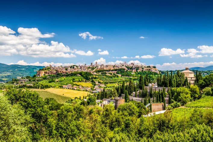 Stadt Orvieto in Umbrien auf einem Hügel, umgeben von grünen Feldern und Zypressen im Vordergrund; blauer Himmel mit weißen Wolken im Hintergrund.