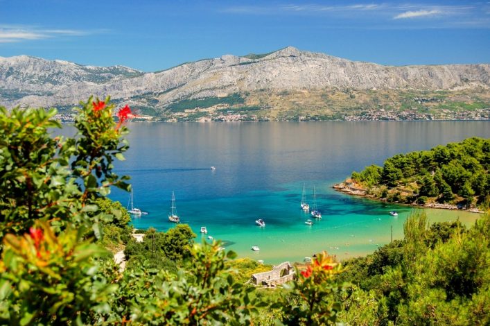 Klares, türkisblaues Wasser an der Lovrecina-Strandbucht auf Brac, Kroatien, mit dichtem Grün im Vordergrund, Booten auf dem Wasser in der Mitte und majestätischen Bergen im Hintergrund.