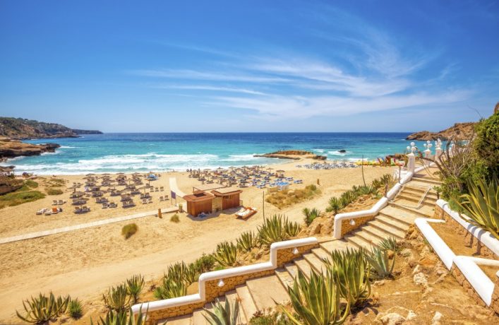 Breiter Sandstrand mit Liegestühlen und Sonnenschirmen, umgeben von klarem, türkisblauem Meer. Rechts führt eine Treppe mit Geländer zur Strandpromenade hinab, flankiert von grünen Pflanzen.