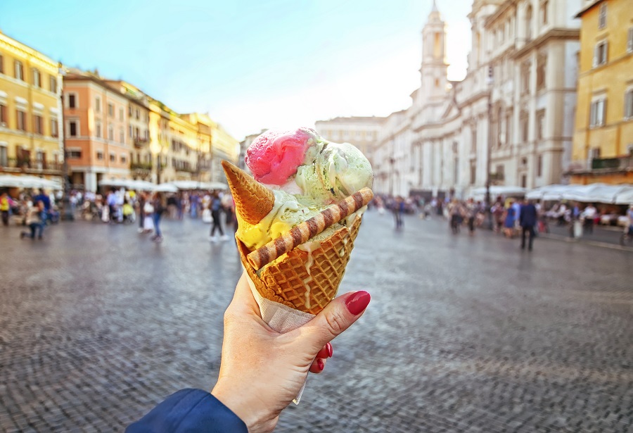 Eine Hand hält ein Eis mit mehreren Sorten in einem Hörnchen, dekoriert mit einer Waffelrolle im Vordergrund. Im Hintergrund befinden sich unscharfe Gebäude und Menschen auf einem Platz.
