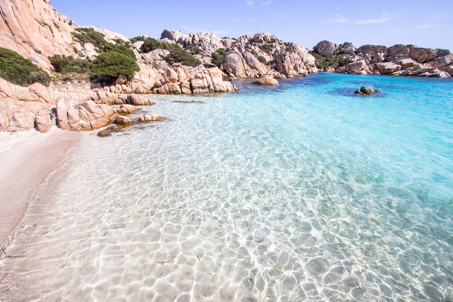 Kristallklares, türkisblaues Meer umspült sanft den weißen Sandstrand Sardiniens, ideal für einen Strandurlaub im August. Pinkfarbene Felsformationen rahmen die Bucht.