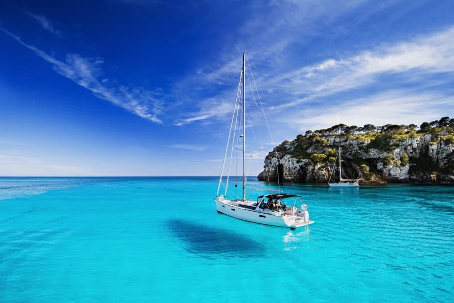 Ein weisses Segelboot schwimmt auf türkisblauem Wasser in einer ruhigen Bucht von Mallorca, umgeben von Klippen und grüner Vegetation. Ideal für einen Familienurlaub auf Mallorca. Im Hintergrund ein weiteres Boot.