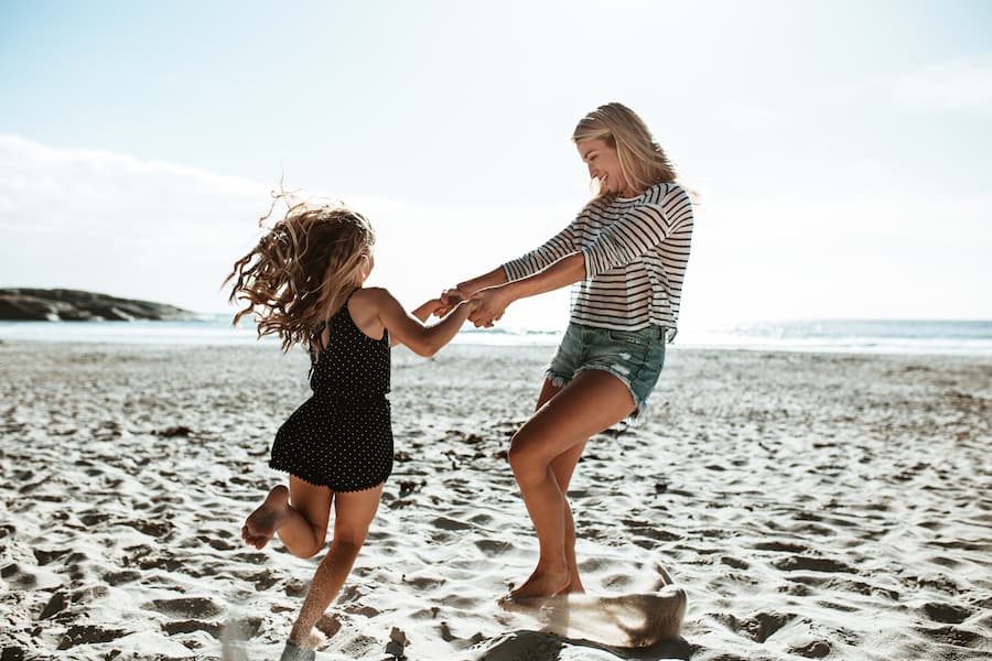 Eine Mutter und ihre Tochter spielen lachend am Strand, halten sich an den Händen und drehen sich im Kreis. Im Hintergrund sieht man das Meer und die klare, helle Himmel. Familienurlaub am Meer.