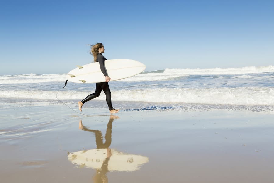Teenager-Mädchen im Neoprenanzug läuft barfuß am Strand entlang und trägt ein Surfbrett unter dem Arm. Familienurlaub am Meer mit sanften Wellen im Hintergrund, unter blauem Himmel.