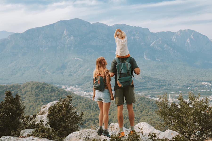 Ein Paar mit Rucksäcken steht auf einem Felsen, den Blick auf bewaldete Berge gerichtet. Der Vater trägt ein kleines Kind auf den Schultern. Perfekter Familienurlaub in der Natur.