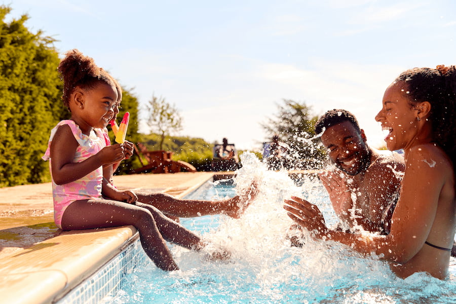 Kind sitzt am Rand eines Pools, hält ein Eis und spritzt lachend seine Eltern nass, die im Wasser sitzen. Die Sonne scheint, Bäume im Hintergrund. Fröhliche Szene eines Sommer-Familienurlaubs.