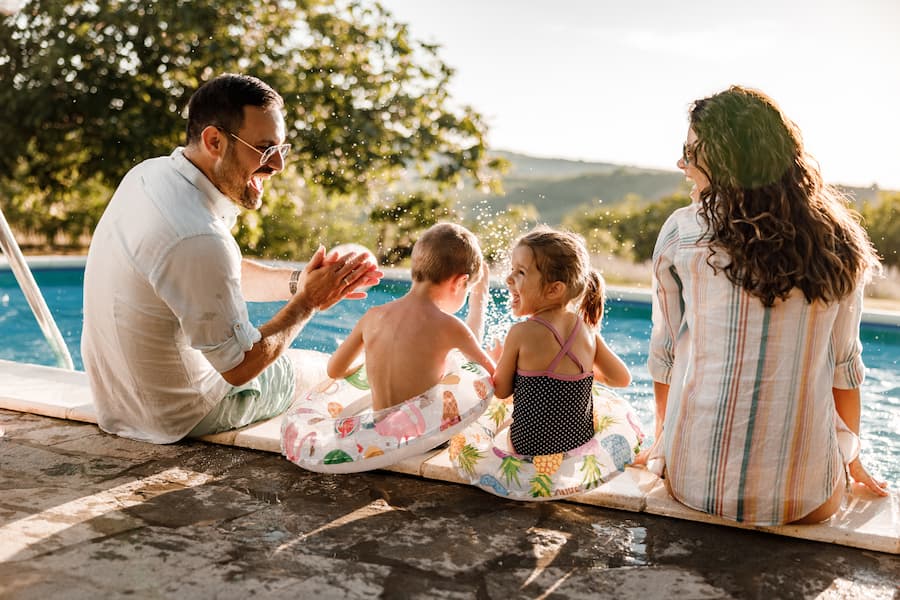Ein Mann, eine Frau und zwei lachende Kinder mit Schwimmreifen sitzen am Rand eines Pools und planschen im Wasser. Im Hintergrund sind Bäume zu sehen. Familienurlaub bei sonnigem Wetter.