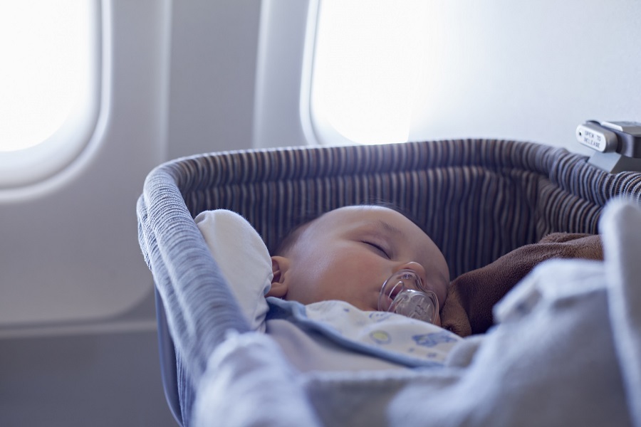 Ein Baby schläft friedlich in einem blauen Reisebettchen mit Schnuller im Mund. Durch die Fenster im Hintergrund sind die typischen Flugzeugfenster zu sehen. Perfekter Moment für einen Familienurlaub.