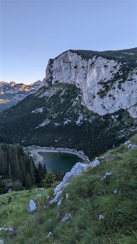 Grüne Wiese führt zu bewaldetem Tal mit dunkelgrünem See. Im Hintergrund erhebt sich eine steile Felswand, die von links nach rechts verläuft. Darüber zeigt sich ein heller, klarer Himmel.