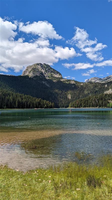 Saphirblauer Gebirgssee umgeben von einem dichten, dunklen Nadelwald; im Hintergrund erhebt sich ein massiver, felsiger Berg unter einem strahlend blauen Himmel mit weißen Wolken; einige Uferpflanzen.