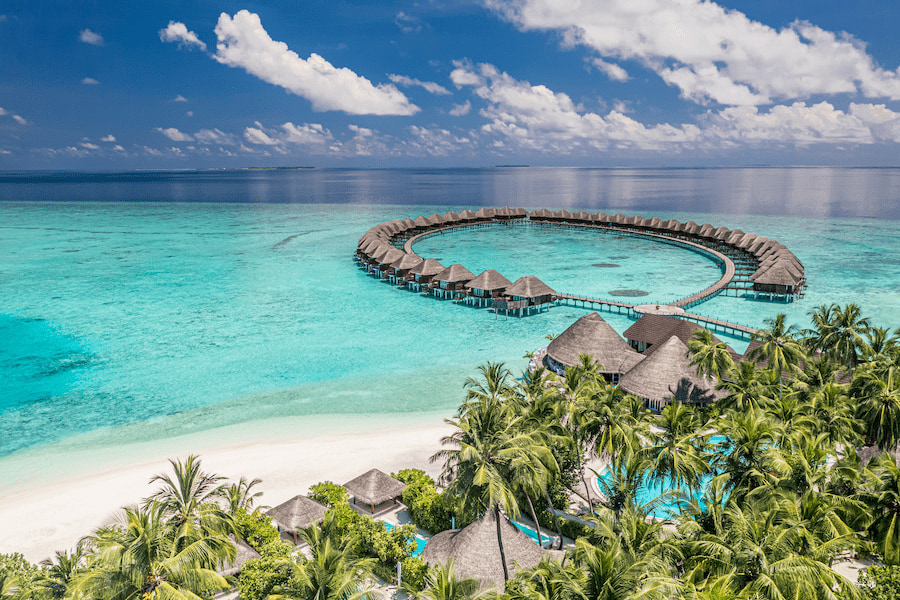 Über türkisblauem Wasser ragen kreisförmig angeordnete Wasserbungalows hinaus. Im Vordergrund erstreckt sich ein weißer Sandstrand, umgeben von Palmen und tropischer Vegetation – ideal für einen Familienurlaub auf den Malediven.