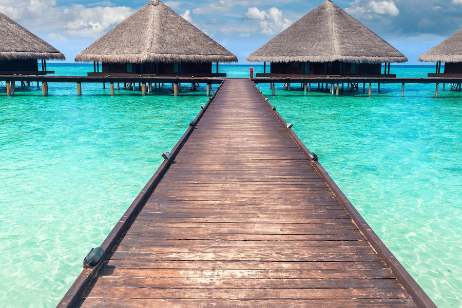 Ein Holzsteg führt zu Wasservillen mit Strohdächern, die auf Pfählen über dem türkisblauen Wasser stehen. Perfekt für einen Familienurlaub auf den Malediven bei sonnigem Klima und klarem Himmel.