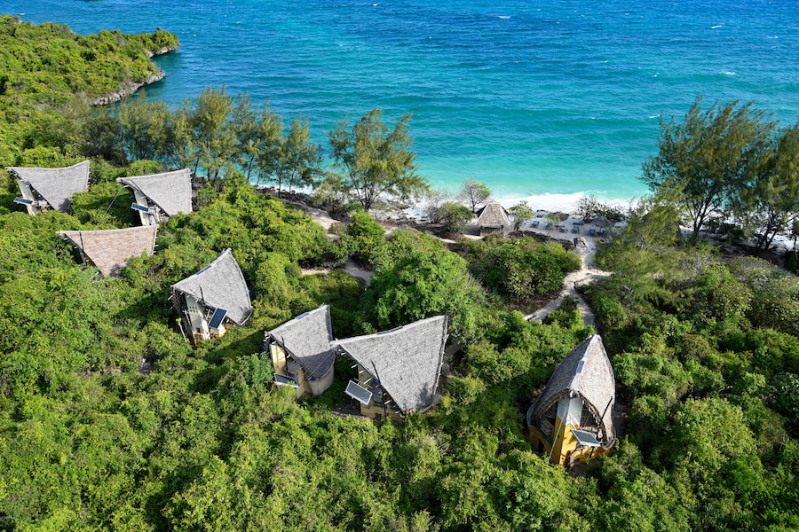 Luftaufnahme von strohgedeckten Bungalows, die in üppiger grüner Vegetation am türkisblauen Meer liegen. Perfektes Setting für einen Familienurlaub auf Sansibar. Ein Pfad führt zur sandigen Küste.