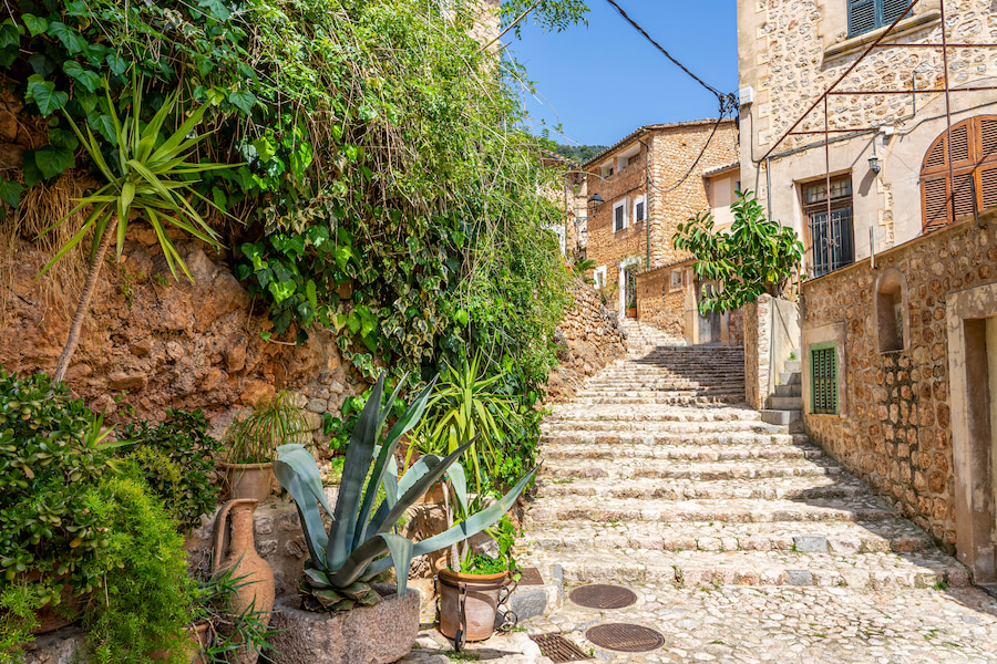 Steintreppe in einem malerischen Dorf auf Mallorca. Links prächtige Grünpflanzen, rechts Häuser aus hellem Naturstein mit grünen Fensterläden. Perfekt für einen Familienurlaub auf Mallorca.