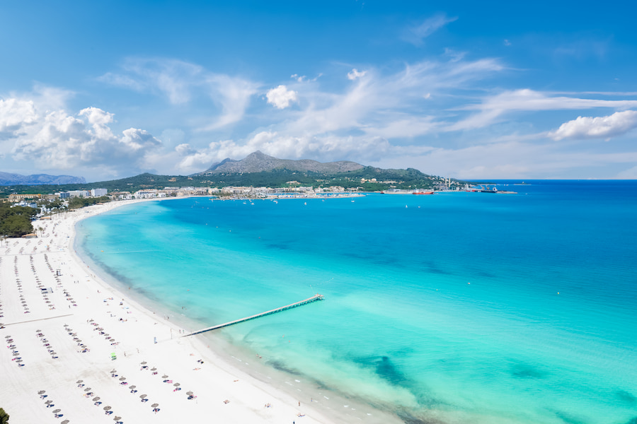 Breiter Sandstrand mit Sonnenschirmen an der Playa de Muro auf Mallorca, ideal für Familienurlaub. Links Hotels und Grünflächen, rechts türkisblaues Meer, im Hintergrund bewaldete Hügel.