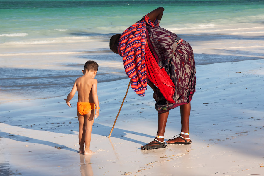 Ein Massai mit buntem Gewand und einem Stock zeigt einem kleinen Jungen im orangefarbenen Badeanzug etwas im Sand am Strand von Sansibar. Ideal für Familienferien auf Sansibar.