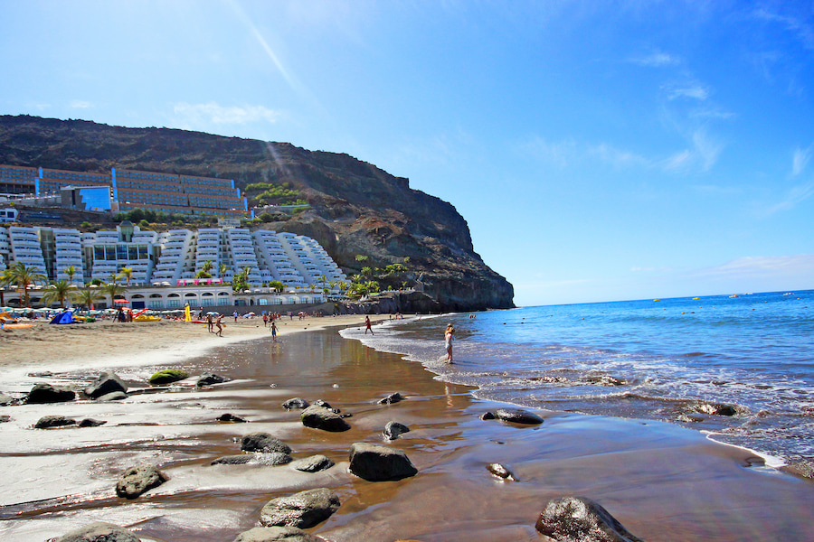 Sonniger Strand von Taurito, links Palmen und Hotelanlagen, rechts das blaue Meer. Im Vordergrund Familien am Wasser, ideal für Familienurlaub auf Gran Canaria. Felsen im Sand verstreut.