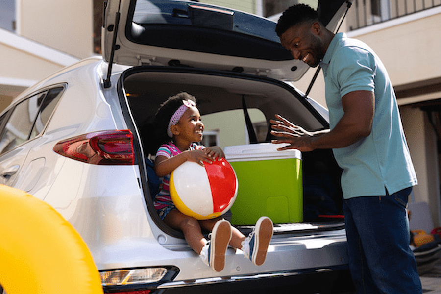 Vater und Tochter packen das Auto für den Familienurlaub auf Gran Canaria. Die Tochter sitzt im Kofferraum mit einem Ball, der Vater steht lächelnd daneben. Eine Kühlbox ist ebenfalls im Kofferraum.