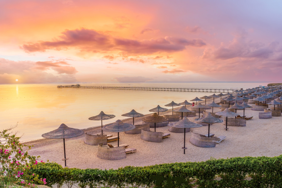 Sonnenuntergang am goldenen Strand von Marsa Alam. Zahlreiche Strohhüte und Liegen stehen auf dem Sand, während das Meer sanft die Küste umspült. Ein langer Steg erstreckt sich ins Wasser.