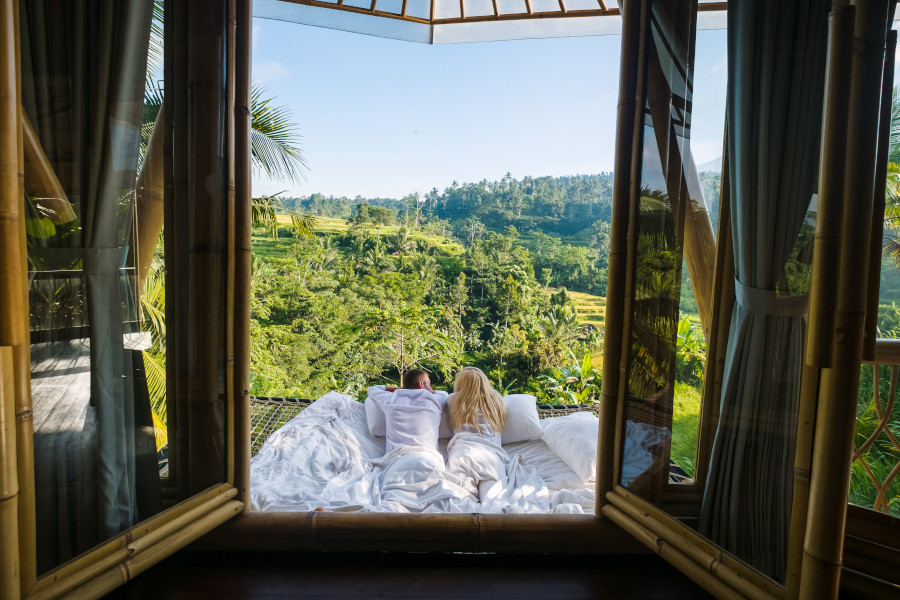 Ein Paar liegt in einem Bett aus weißen Laken in einem offenen Bambuszimmer mit Blick auf eine grüne, hügelige Landschaft. Palmen umrahmen die Szenerie unter einem klaren Himmel.