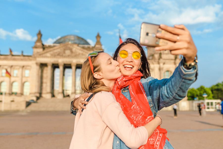 Zwei Frauen in Umarmung machen ein Selfie vor dem Reichstagsgebäude in Berlin. Die rechte Frau trägt eine Sonnenbrille und ein rotes Tuch, die linke küsst sie auf die Wange.