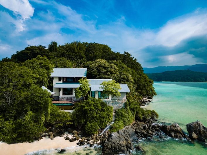 Luxuriöses Hotel auf den Seychellen direkt am türkisfarbenen Meer, eingebettet in üppige grüne Vegetation. Im Vordergrund eine kleine Sandbucht und felsige Küste, darüber blauer Himmel mit Wolken.