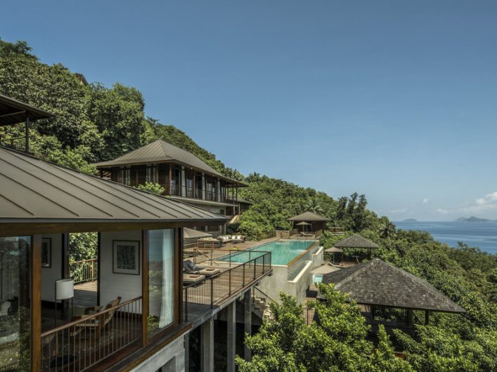 Luxuriöses Resort am Hang, umgeben von üppigem Grün, mit mehreren Gebäuden im traditionellen Stil. Zentraler langer Infinity-Pool bietet atemberaubenden Meerblick, strahlend blauer Himmel im Hintergrund.