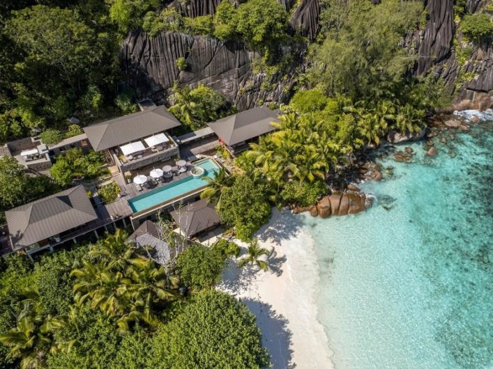 Luxuriöse Strandvillen mit Pool liegen zwischen üppigem Grün und Felsen, daneben ein weißer Sandstrand mit klarem, türkisfarbenem Wasser. Palmen säumen den Rand der Villa, die naturverbunden wirkt.