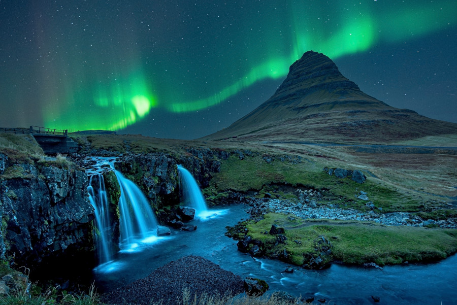 Grüne Nordlichter tanzen über dem nächtlichen Himmel. Im Vordergrund fließt ein Wasserfall über Felsen. Rechts erhebt sich ein markanter, grasbedeckter Berg unter einem Sternenhimmel.