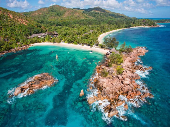 Türkises Wasser umgibt felsige Inselgruppe mit üppiger Vegetation. Ein Segelboot links im Wasser. Im Hintergrund dichter Wald und geschwungener Sandstrand unter blauem Himmel.